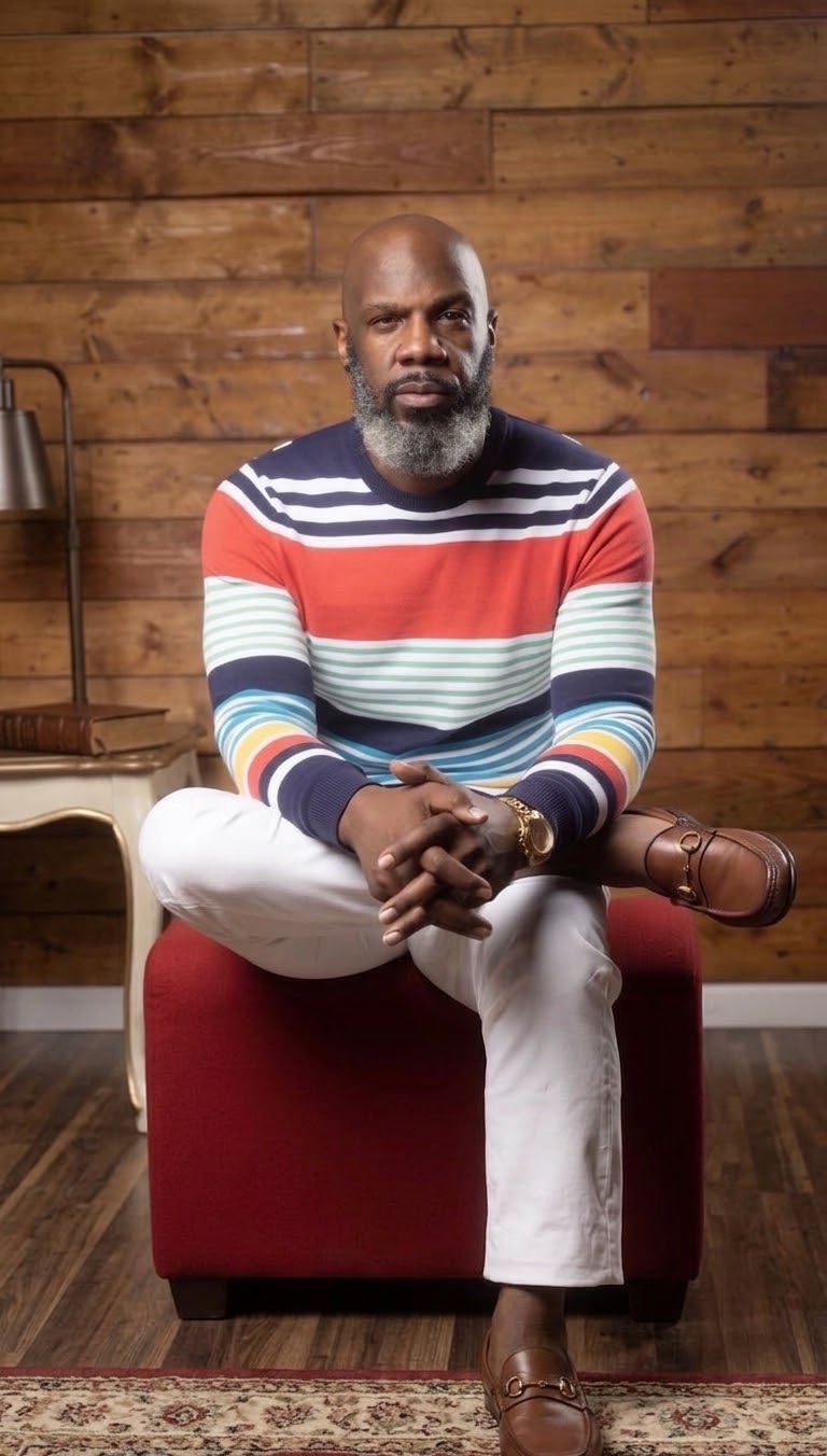 Mr. Robinson sitting on a red chair. He is sitting in front of a wood paneled wall with a striped long-sleeved shirt and white pants.