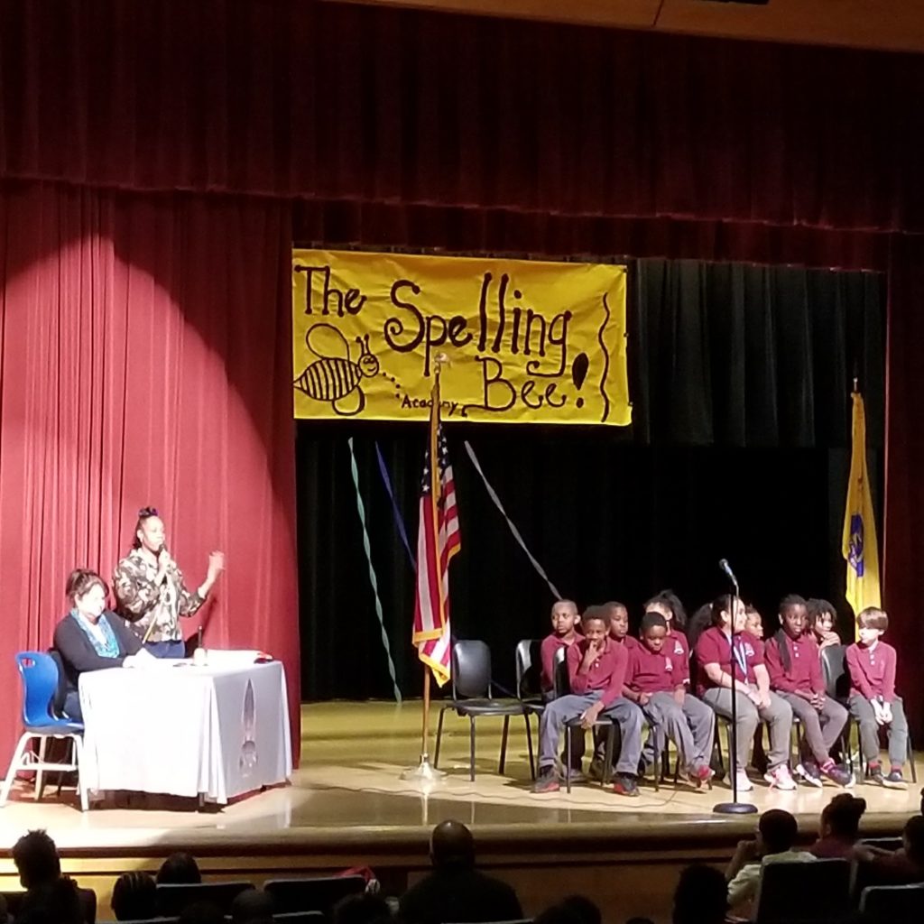 Spelling Bee November 2017; students on stage left; judges on stage right. All brightly lit. Yellow sign in background says "The Spelling Bee" with a picture of a bee.
